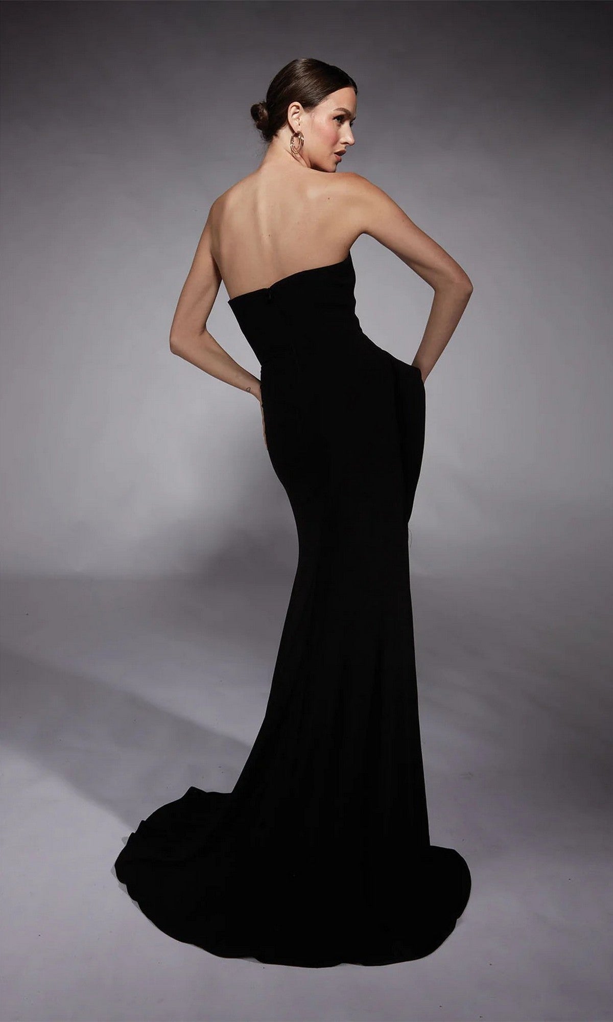 A woman in a strapless formal dress poses with her back to the camera, highlighting the elegant silhouette of her floor-length white Alyce 7112 dress in a studio setting.